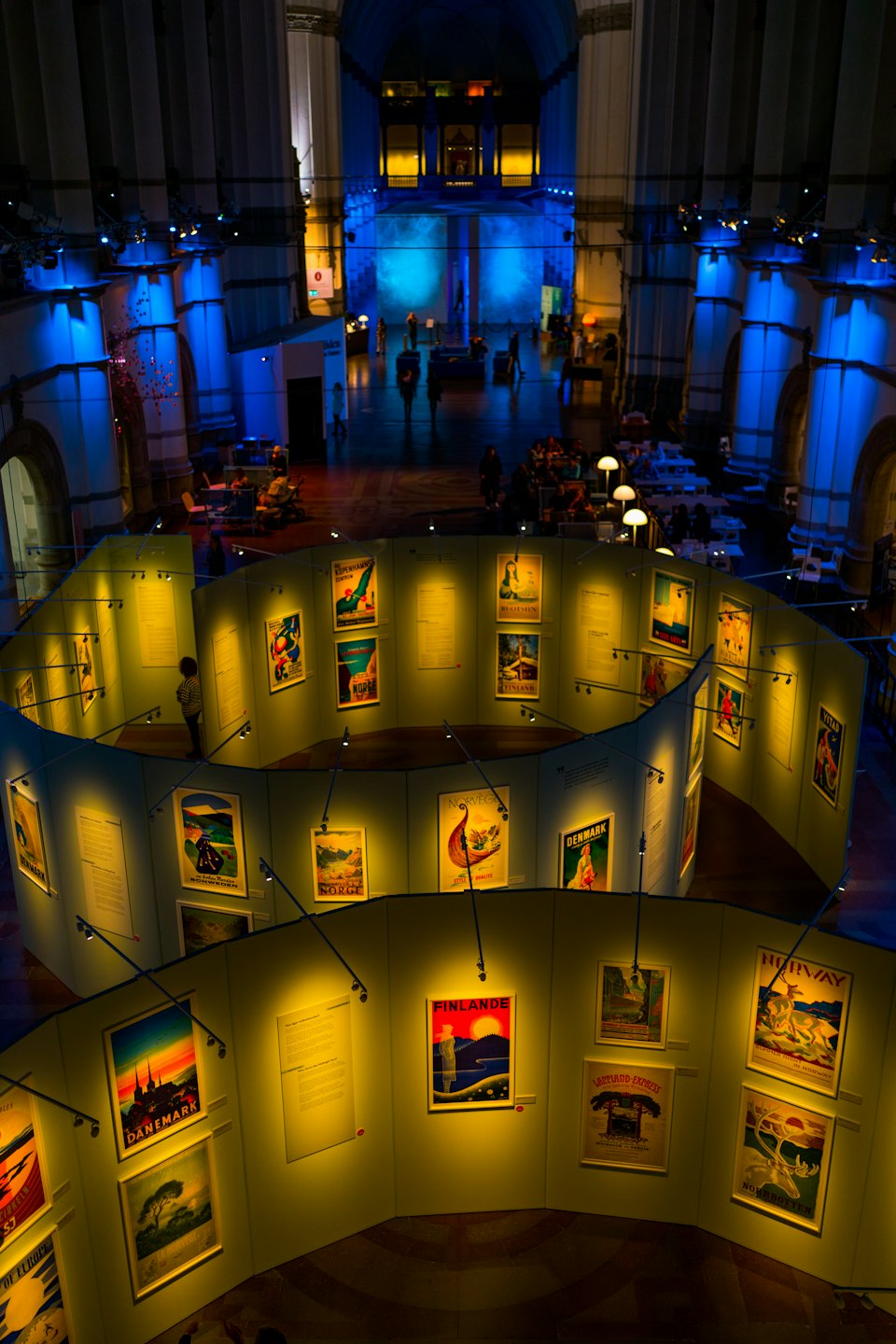 Des visiteurs parcourent une exposition d'affiches de voyage vintage dans un grand hall.